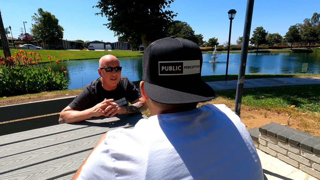 two guys at a bench near a pond talking with the nearest man wearing a public perception brand cap